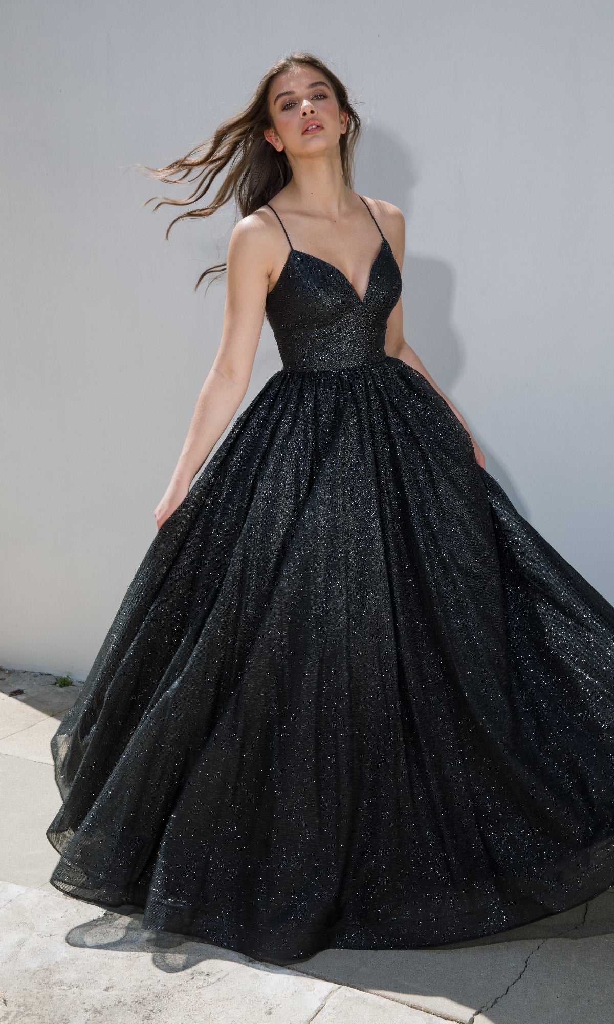 Outdoors, a woman wears the Strappy-Back Glitter Prom Ball Gown: Eureka 9009—a sleeveless, floor-length black A-line dress with thin straps and shimmering fabric, ideal as an evening gown or for special occasions.